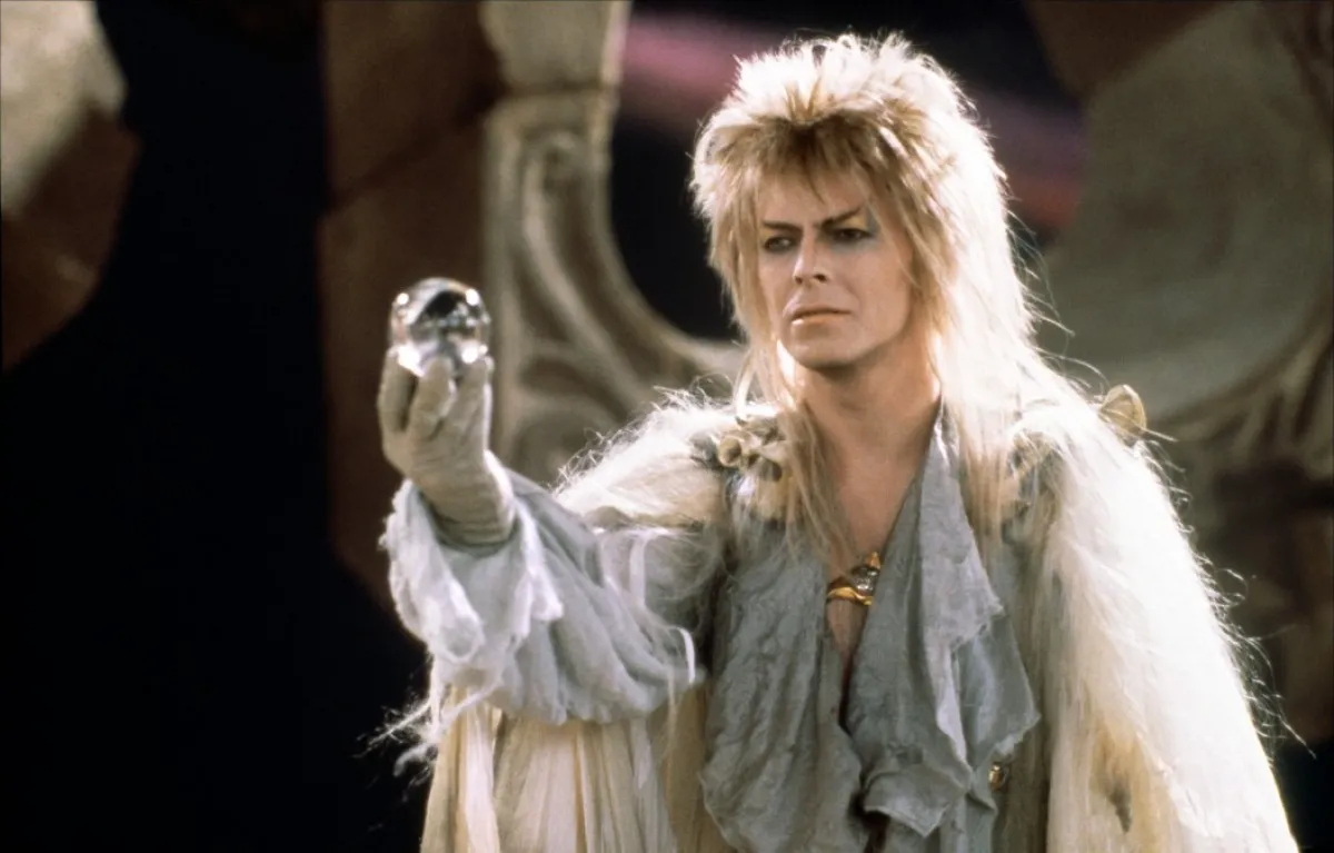 Jareth (David Bowie) holds up a glass orb, wearing a shimmery silver outfit and heavy makeup.