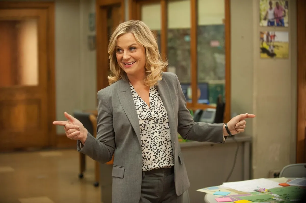 Leslie Knope smiling and making finger guns pointing off camera in Parks and Recreation.