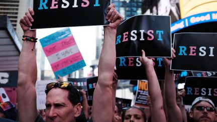 Protesters hold signs reading RESIST, written in blue, pink and white, the colors of the transgender flag.