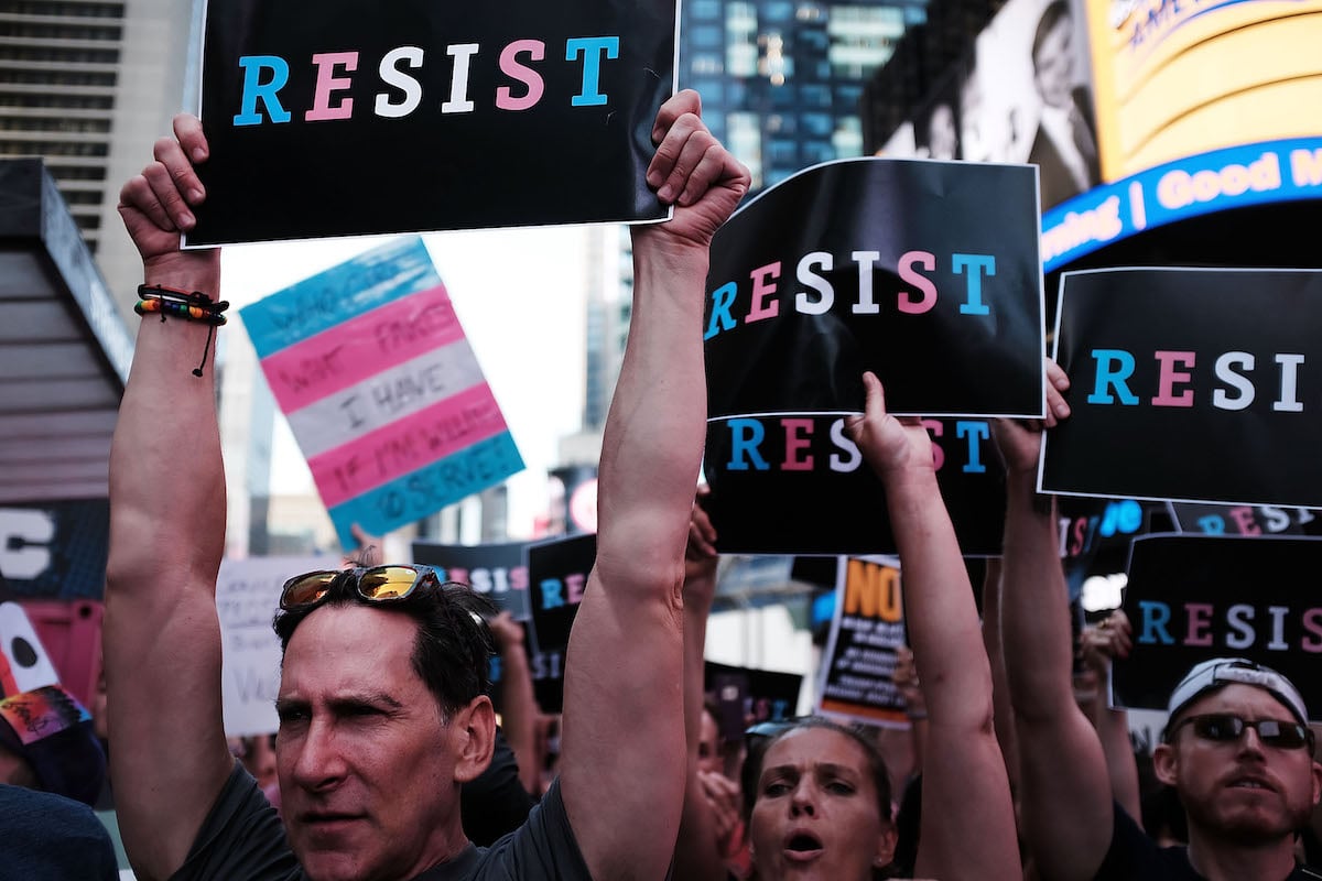 Protesters hold signs reading RESIST, written in blue, pink and white, the colors of the transgender flag.