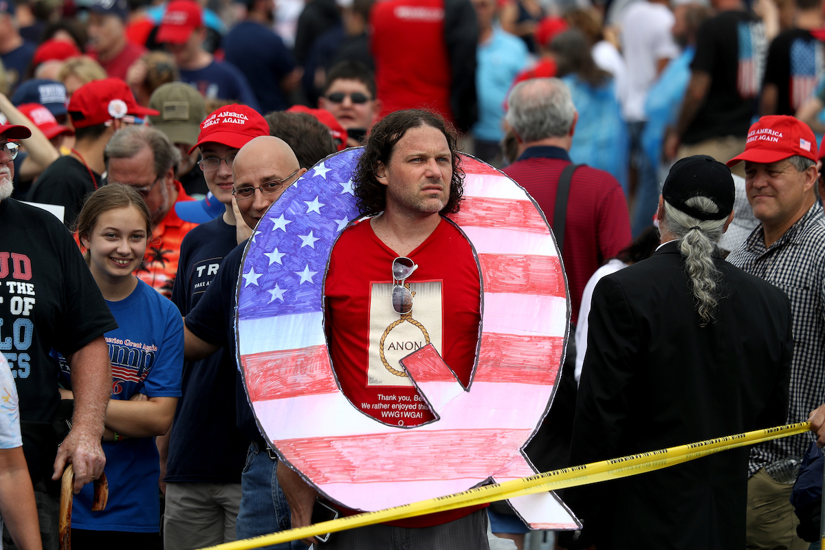 Trump supporter with a giant Q-shaped sign hanging around his neck looks sad and tired.