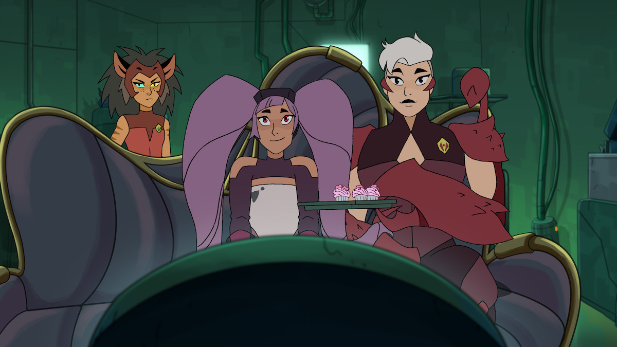 She-Ra's anti-heroes, Catra, Entrapta, and Scorpia, sit on a sofa eating cupcakes.