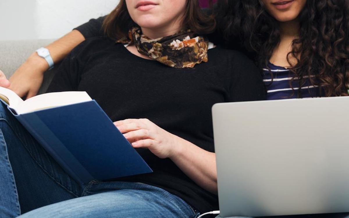 Couple lounging together, one woman holding a book and the other with a computer on her lap.