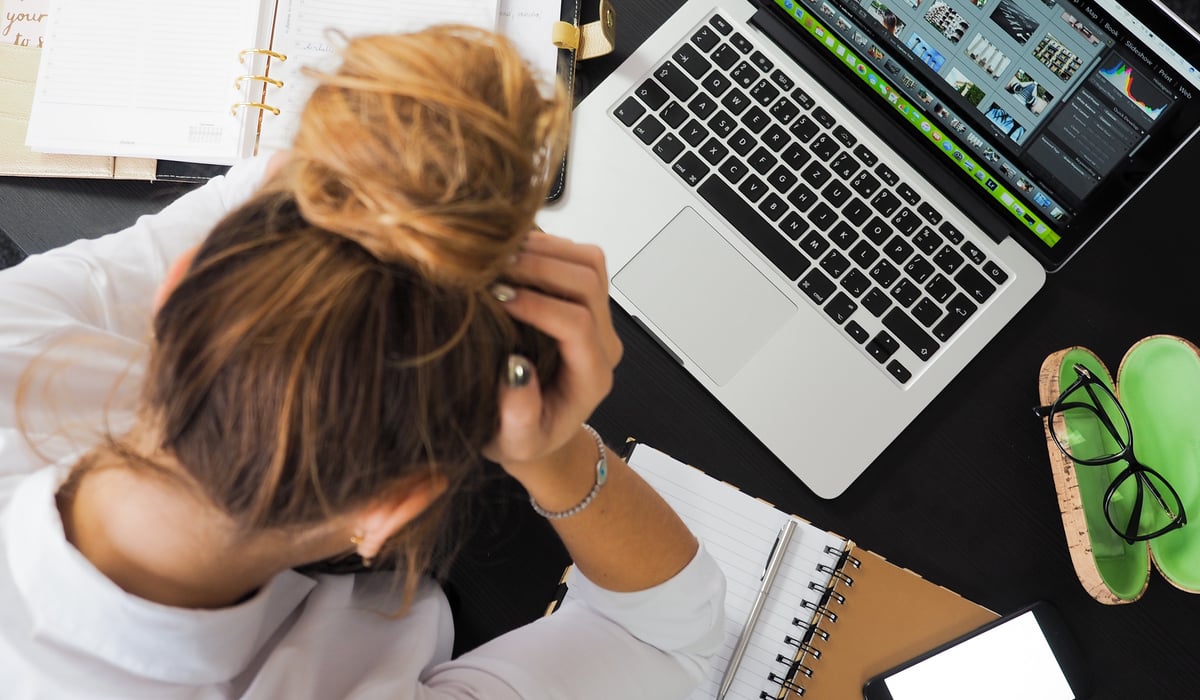 A woman, frustrated, holding her head in her hands on her desk above her laptop.