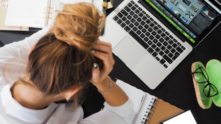 A woman, frustrated, holding her head in her hands on her desk above her laptop.