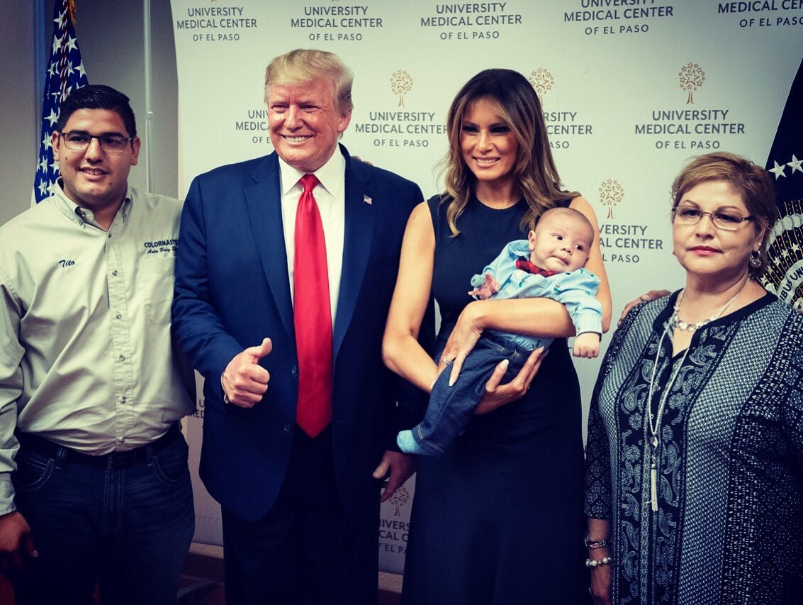 Melania Trump holds an infant for photos while Donald grins and gives a thumbs-up. 