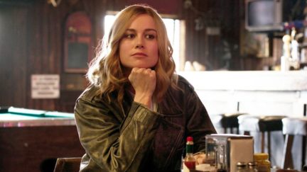 Captain Marvel sits with her head in her chin and a smirk.