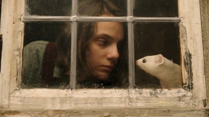 Lyra (Dafne Keen) looks at her daemon, a white ferret named Pantalaimon, behind a window in a scene from 'His Dark Materials.'