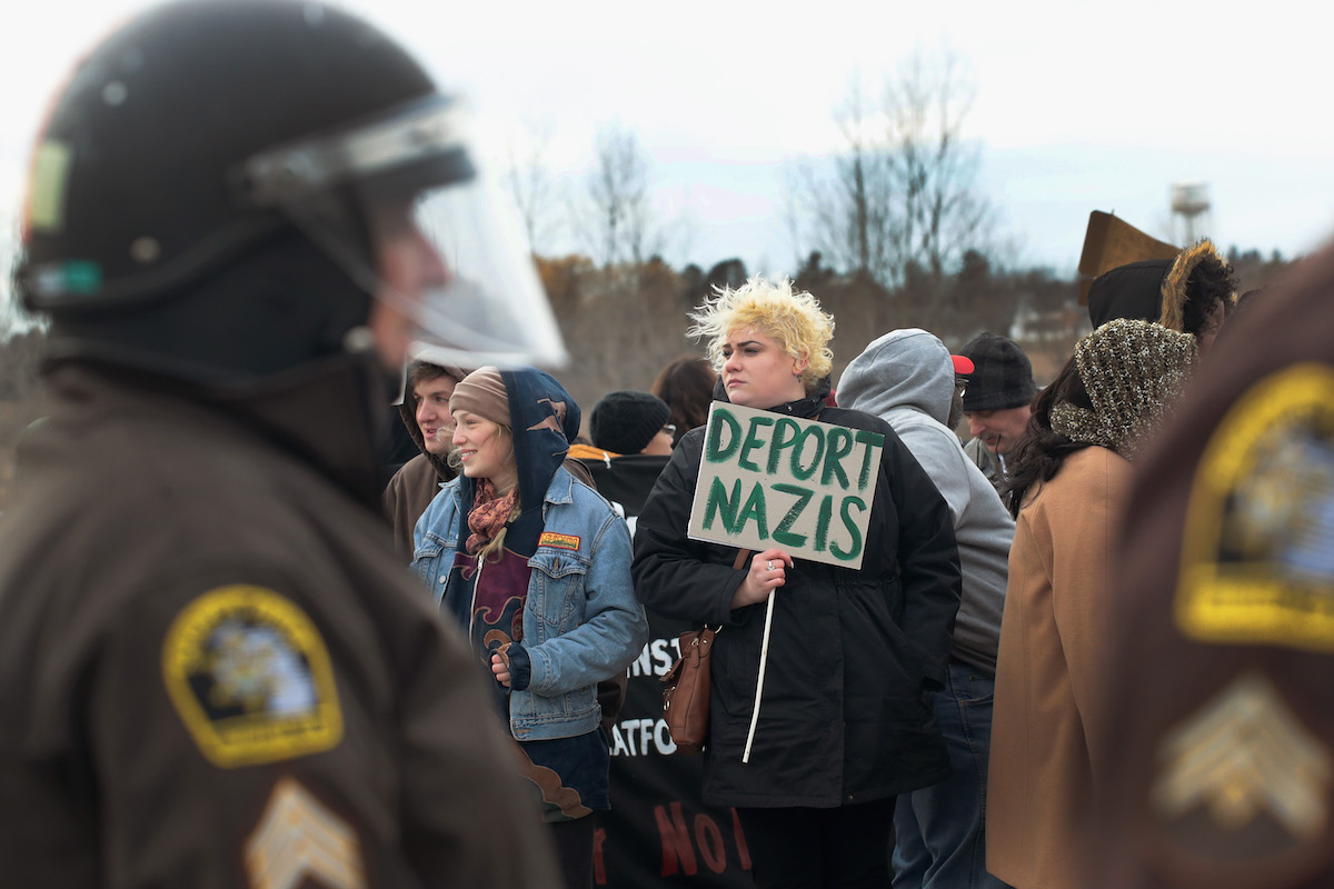 Anti-Nazi protester holds a sign saying "Deport Nazis" with police in the foreground.