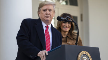 Donald Trump stands at a podium, in front of Melania, looking smugly at the camera.