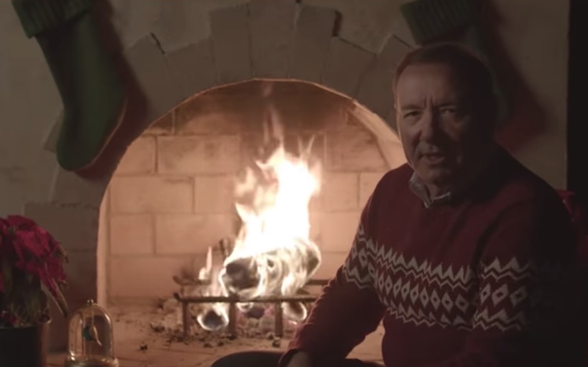 Kevin Spacey as Frank Underwood sits in front of a fireplace in a Christmas sweater, looking into the camera.