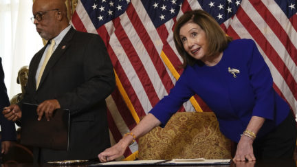 Nancy Pelosi leans over a desk in front of US flags.
