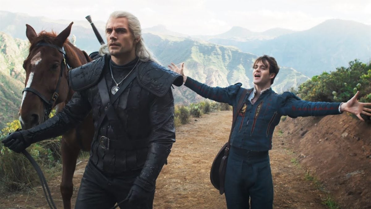Geralt walks along with his horse while Jaskier gestures enthusiastically with arms wide in Netflix's The Witcher.