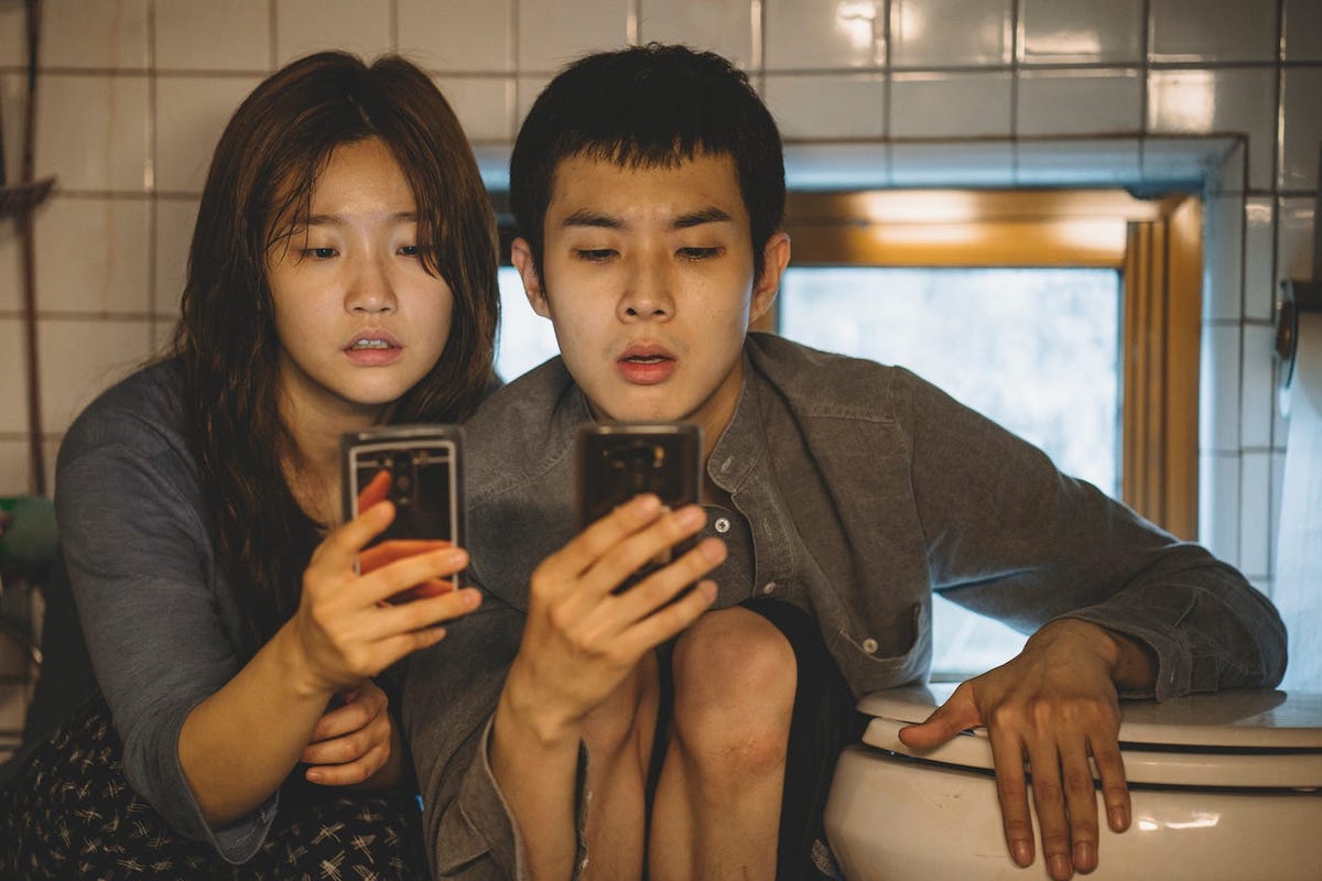Characters from Parasite look at their phones in front of a toilet in their semi-basement apartment.