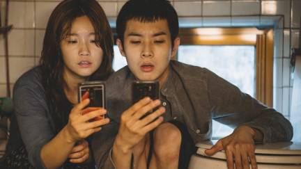 Characters from Parasite look at their phones in front of a toilet in their semi-basement apartment.