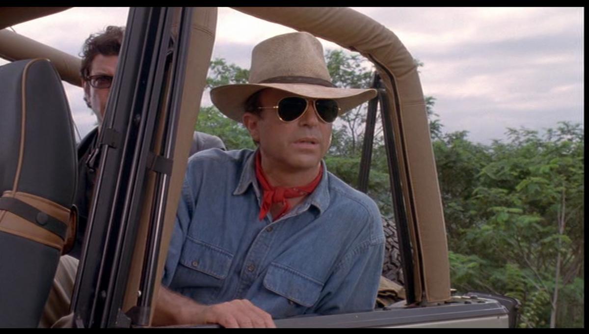 A white man wearing a wide fedora and sunglasses sitting in a jeep looking into the distance in "Jurassic Park"
