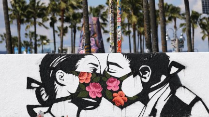 Palm trees stand behind a street art piece by artist Pony Wave depicting two people kissing while wearing face masks on Venice Beach on March 21, 2020 in Venice, California.