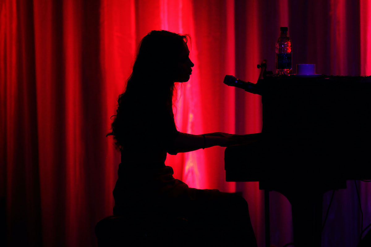 Fiona Apple plays piano in silhouette against a red curtain.