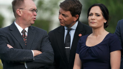Stephanie Grisham stares at the camera while two men talk