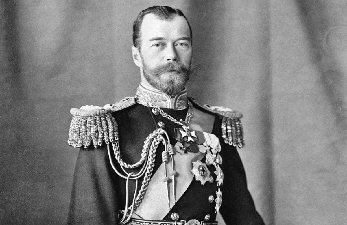 Gelatin silver print photograph of Nicholas II, Emperor of Russia. He is standing with his left hand resting on a bicorn hat on the decorative table beside him to the right. He is wearing an ornate naval uniform including epaulets, a sash and insignia. There is a pair of white gloves in his right hand and a sword by his side.