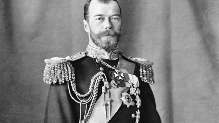 Gelatin silver print photograph of Nicholas II, Emperor of Russia. He is standing with his left hand resting on a bicorn hat on the decorative table beside him to the right. He is wearing an ornate naval uniform including epaulets, a sash and insignia. There is a pair of white gloves in his right hand and a sword by his side.