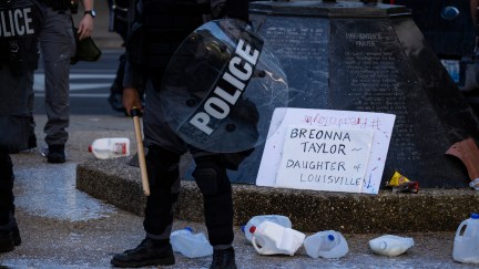 Police officers in riot gear stand in and around milk jugs and a sign honoring Breonna Taylor.