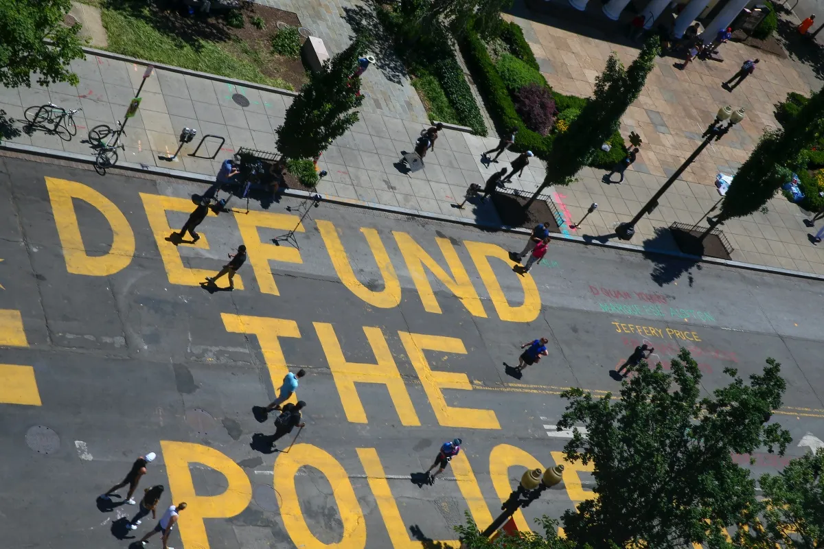 "Defund the Police" is written in large yellow lettering on a city street, seen from above.
