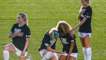 Women's soccer players take a knee and cry on the field while one player stands.