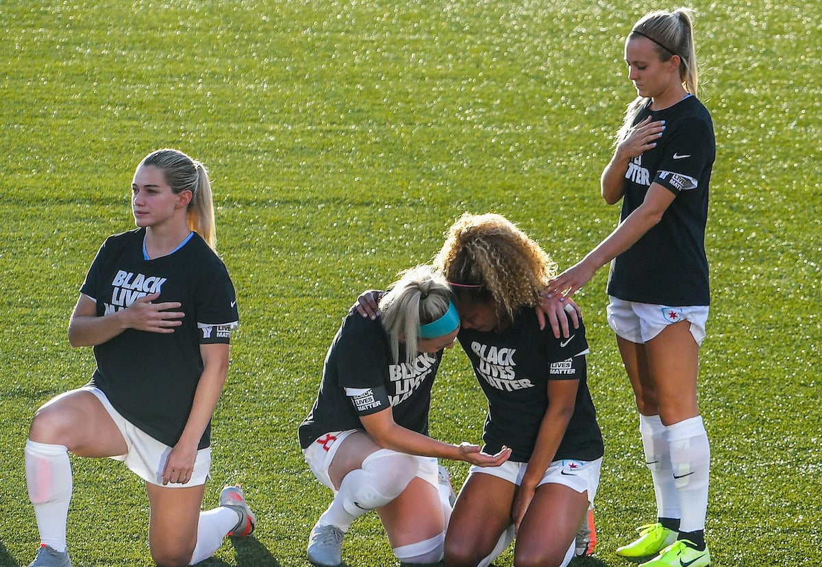 Women's soccer players take a knee and cry on the field while one player stands.