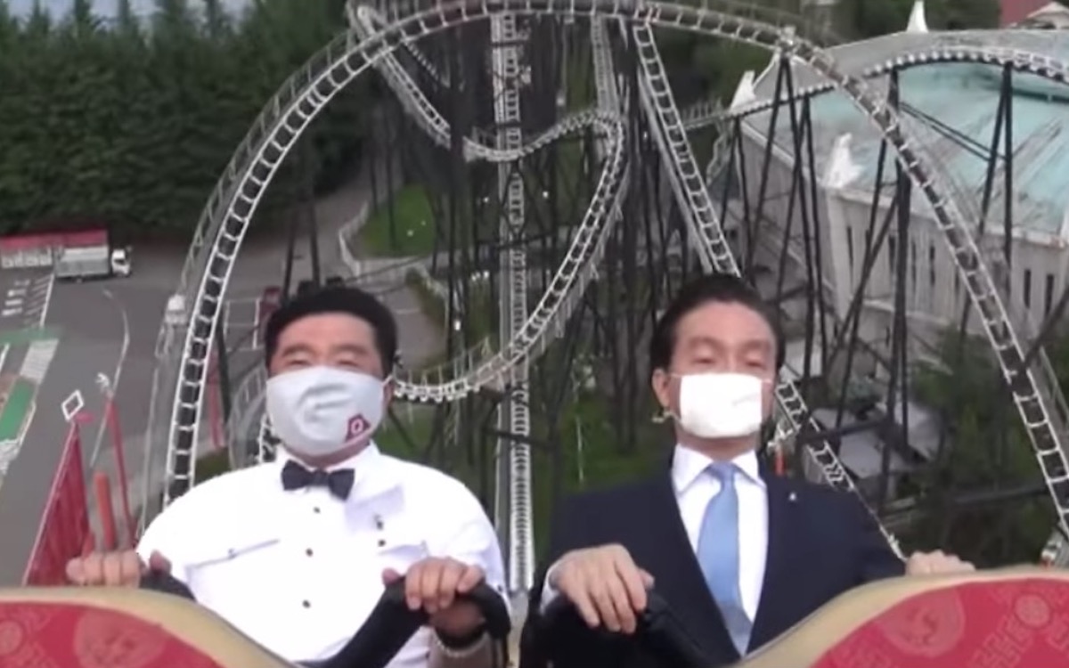 Two Japanese executives ride a rollercoaster wearing masks, expressionless.