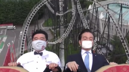 Two Japanese executives ride a rollercoaster wearing masks, expressionless.