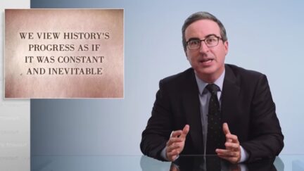 John Oliver sits at his desk with a placard reading 