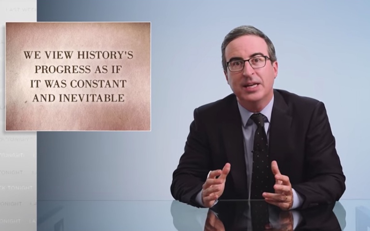 John Oliver sits at his desk with a placard reading "we view history's progress as if it was constant and inevitable."