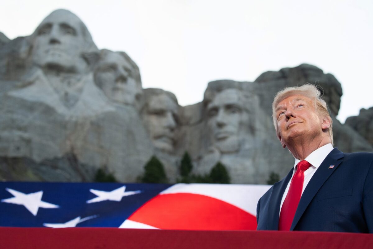 trumpstands in front of an image of mt. rushmore, smiling vacuously