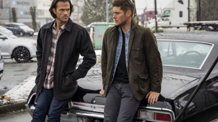 Sam and Dean lean on a car outside while having a conversation in 