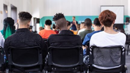 Students sit in a classroom setting, facing a teacher speaking.