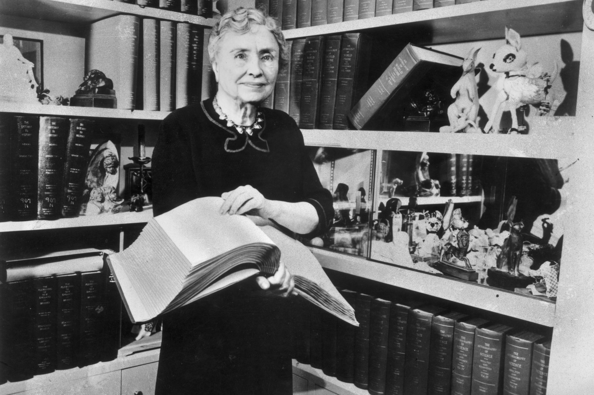 1956: Portrait of American writer, educator and advocate for the disabled Helen Keller (1880 - 1968) holding a Braille volume and surrounded by shelves containing books and decorative figurines. A childhood illness left Keller blind, deaf and mute. (Photo by Hulton Archive/Getty Images)