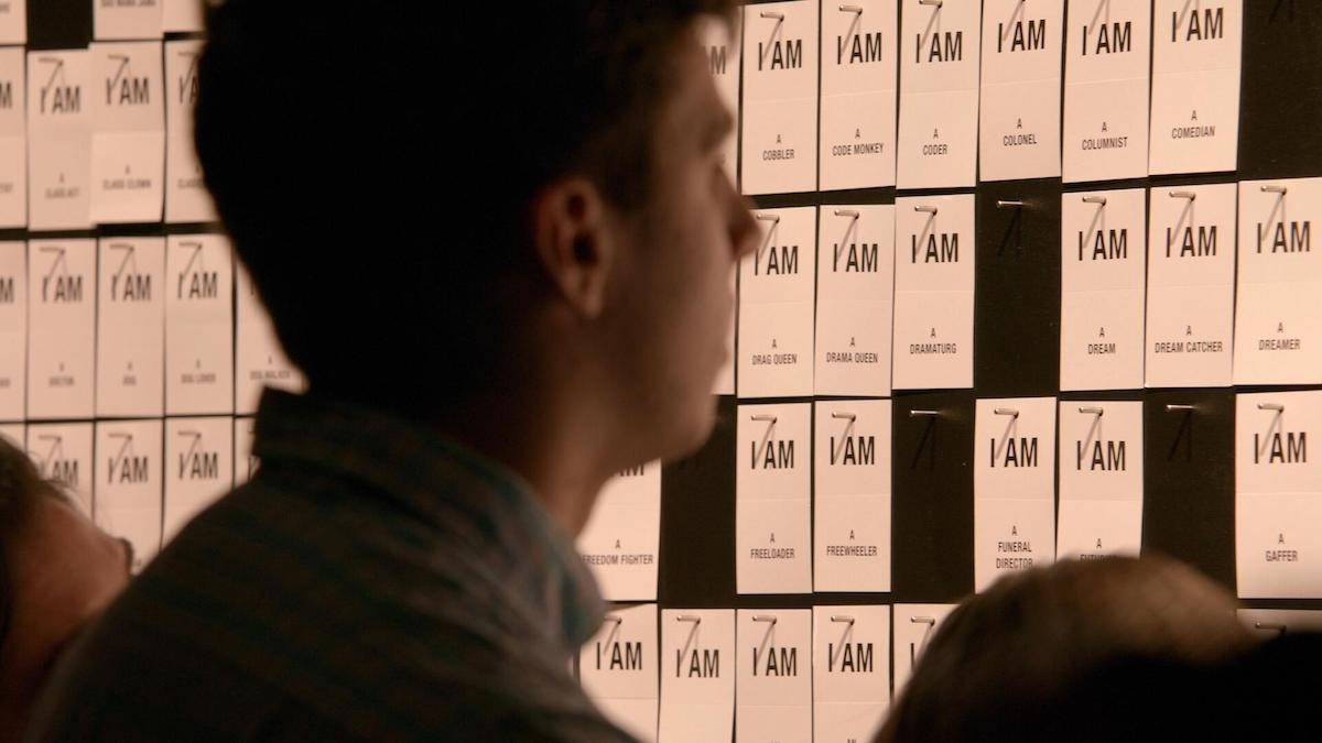 An audience member looks at a wall of cards reading "I AM" with different descriptors on each.