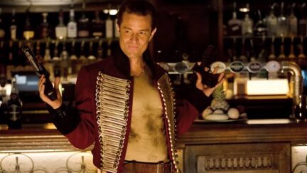 James Marsters as Captain John Hart on Torchwood, holding up two guns and smiling.