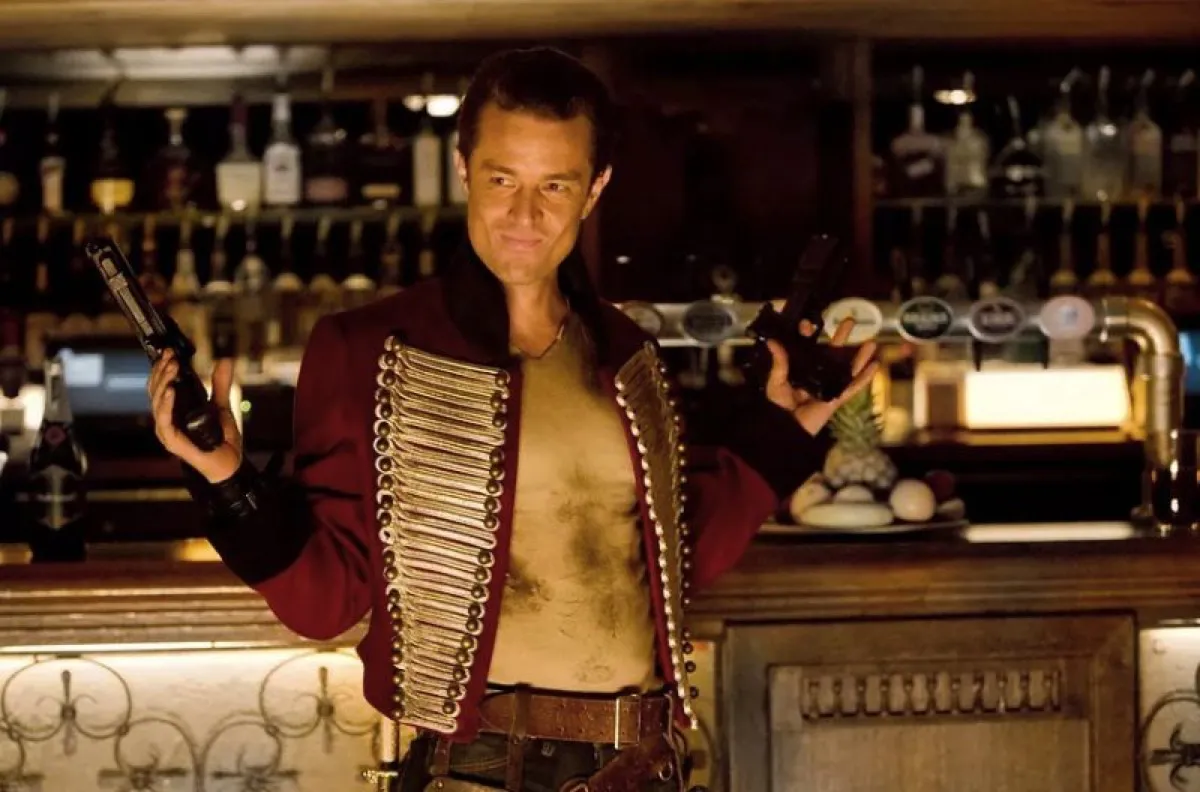 James Marsters as Captain John Hart on Torchwood, holding up two guns and smiling.