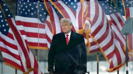 Donald Trump speaks during an outdoor rally, standing in front of American flags.
