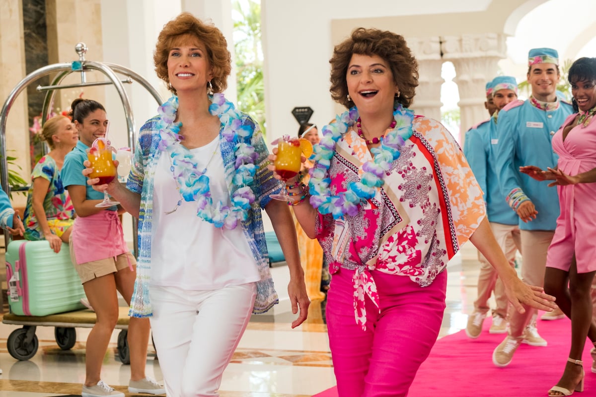 Kristen Wig as Star and Annie Mumolo as Barb in Barb and Star Go to Vista Del Mar, arrive at a brightly colored hotel, carrying tropical drinks.