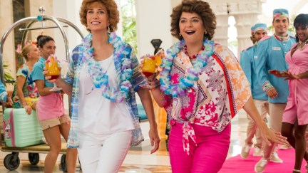Kristen Wig as Star and Annie Mumolo as Barb in Barb and Star Go to Vista Del Mar, arrive at a brightly colored hotel, carrying tropical drinks.
