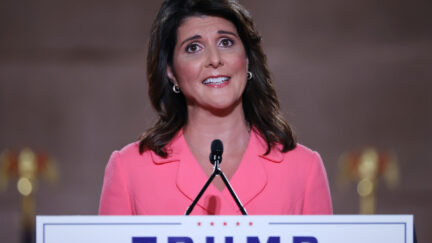 Nikki Haley gives a speech at a podium with a Trump sign.