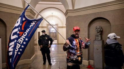 A man wearing a shirt with pictures of Donald Trump on it and carrying a Trump flag walks through the Capitol building and flips off the camera.