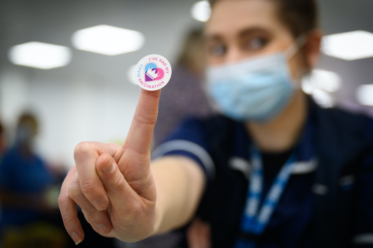 A person in medical gear holds up a sticker reading "I've had my COVID vaccination"
