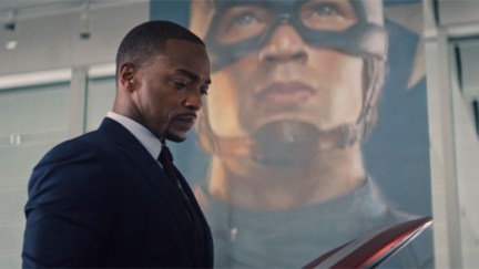 Sam Wilson looks at the Captain America Shield, standing in front of a banner with Steve Rogers' face.