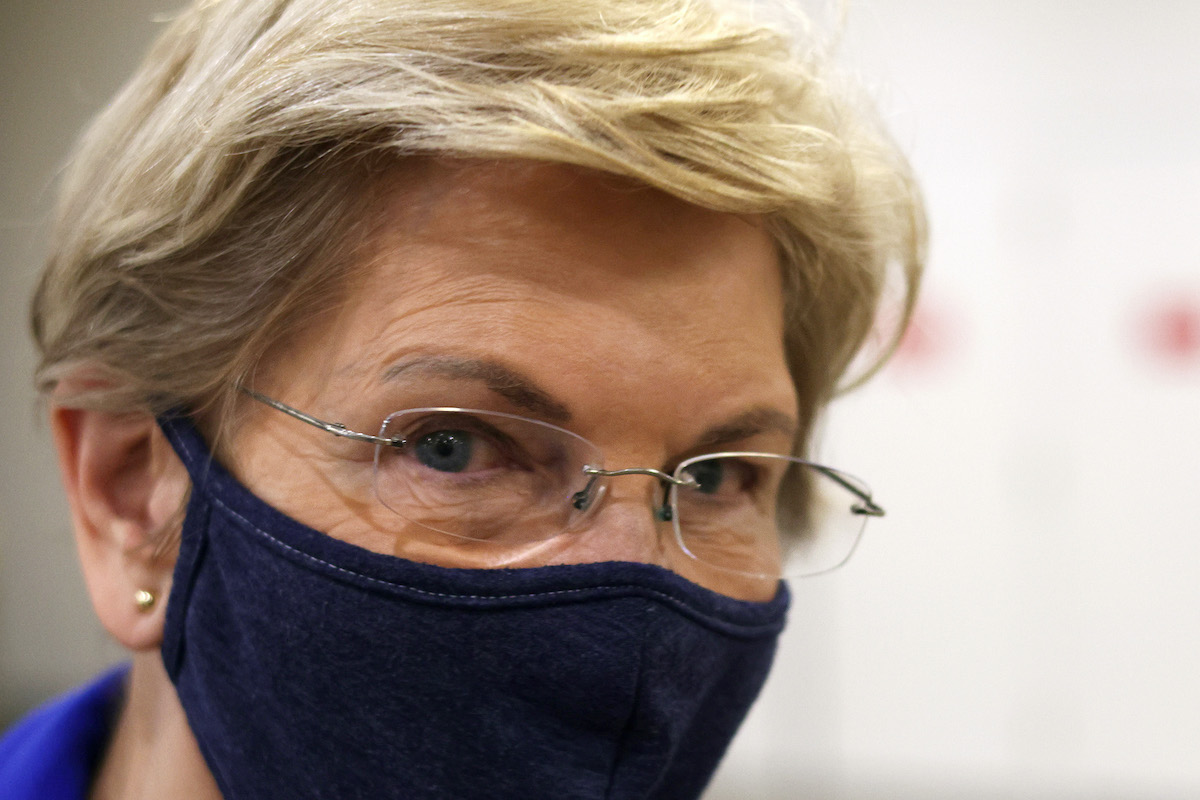 A closeup of Elizabeth Warren, wearing a mask, looking into the camera