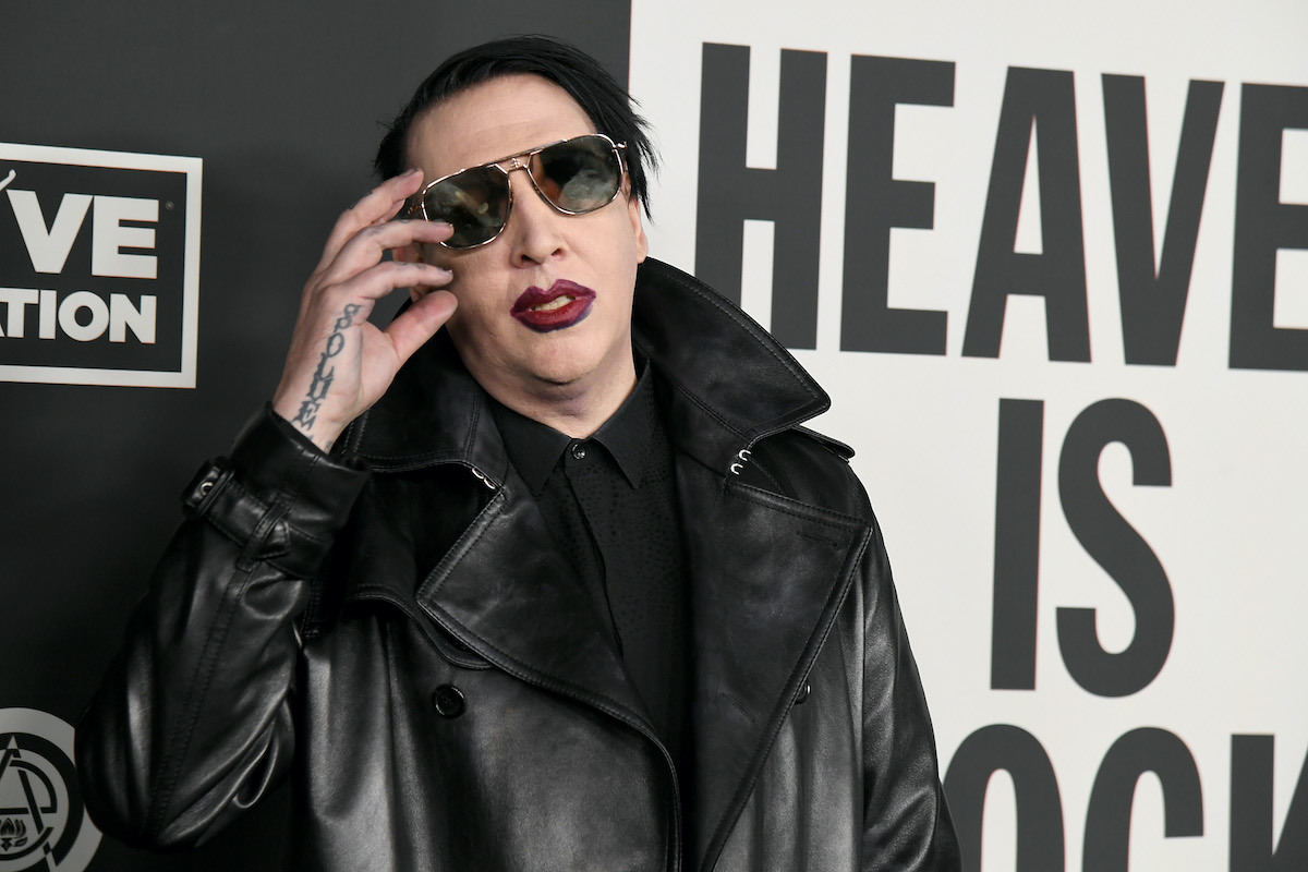 Marilyn Manson wears sunglasses, red lipstick, and a leather jacket in front of a red carpet backdrop reading "Heaven is Rock"