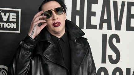 Marilyn Manson wears sunglasses, red lipstick, and a leather jacket in front of a red carpet backdrop reading 